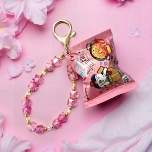 Pode incluir: Um chaveiro rosa com um pacote em miniatura de macarrão instantâneo. O chaveiro tem um fecho dourado e uma alça de contas com contas rosa e douradas. O fundo é rosa com detalhes florais.