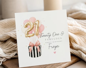 Op de afbeelding: Vierkante verjaardagskaart met de tekst "Twenty One & Fabulous Happy Birthday Freya". De kaart toont een cadeau doos met een roze strik, ballonnen en het gouden cijfer 21.