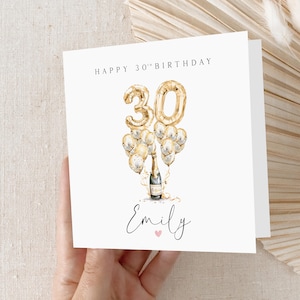 May include: A white card with gold lettering that says "Happy 30th Birthday". There is a gold number 30 balloon with gold and white balloons surrounding it. A bottle of champagne is in the middle of the balloons. The name "Emily" is written in cursive at the bottom of the card.