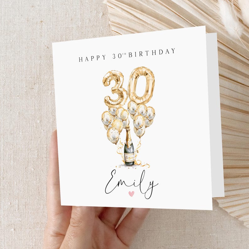 May include: A white card with gold lettering that says "Happy 30th Birthday". There is a gold number 30 balloon with gold and white balloons surrounding it. A bottle of champagne is in the middle of the balloons. The name "Emily" is written in cursive at the bottom of the card.