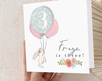 May include: A white card with a blue and pink balloon with the number 3 on it. A white bunny with a flower crown is holding the balloons. The text on the card reads "Freya is three!"
