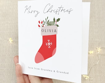 May include: A white Christmas card with a red stocking filled with presents and a candy cane. The stocking has the name Olivia on it and the text "Merry Christmas" and "love from Grandma & Grandad".