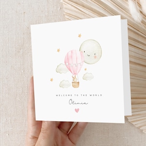 May include: A pink and white hot air balloon with a bunny inside, floating in the sky with a smiling moon and stars. The text "Welcome to the world Olivia" is written below the balloon.
