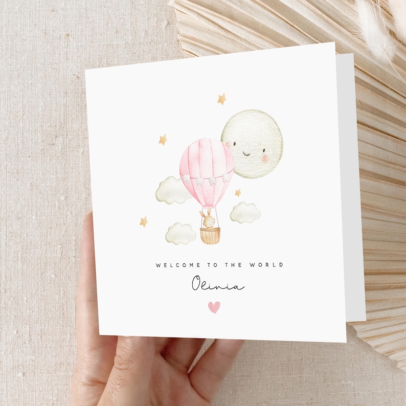 May include: A pink and white hot air balloon with a bunny inside, floating in the sky with a smiling moon and stars. The text "Welcome to the world Olivia" is written below the balloon.
