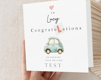 May include: A congratulatory card with a pink heart, the name "Lucy" and the word "Congratulations" with a red "L" sticker. A blue car with a white L sticker is below the text. The text "On passing your driving test" is below the car.