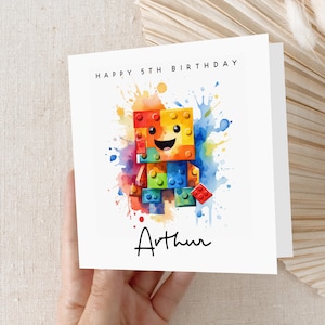 May include: A white card with a colorful watercolor illustration of a smiling Lego figure. The card says "Happy 5th Birthday Arthur".