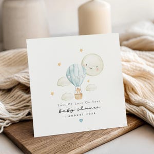 May include: A square greeting card featuring a watercolor illustration of a light blue hot air balloon, a smiling moon, and small yellow stars. The text reads: Lots Of Love On Your baby shower 1 AUGUST 2026, with a small blue heart below the date.