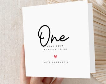 May include: A white card with the word "One" in a black script font. Below, the text reads "Year Down, Forever to Go" and "Love Charlotte" in a smaller, black font. A small pink heart is centered below the text.