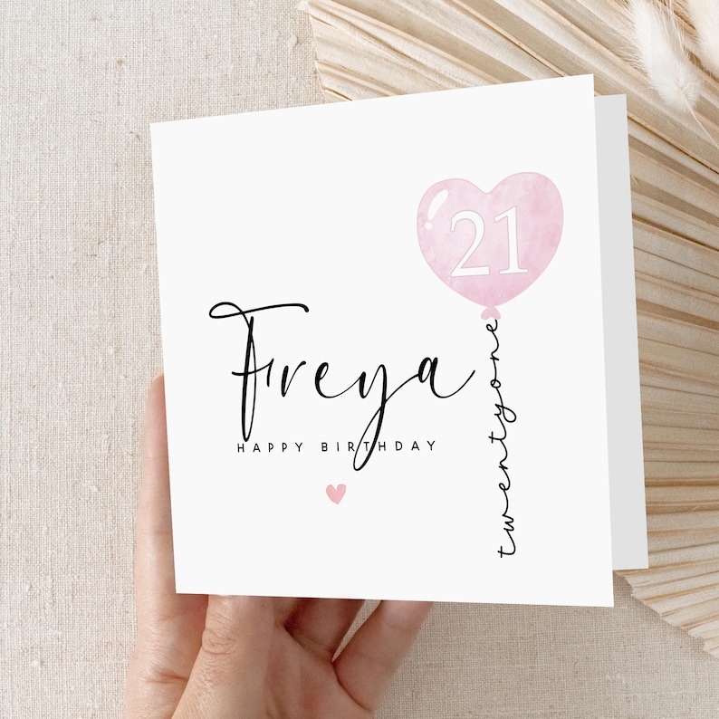 May include: A white birthday card with the name "Freya" in elegant script. A pink heart-shaped balloon with the number "21" is featured. The words "Happy Birthday" and "twentyone" are also present.