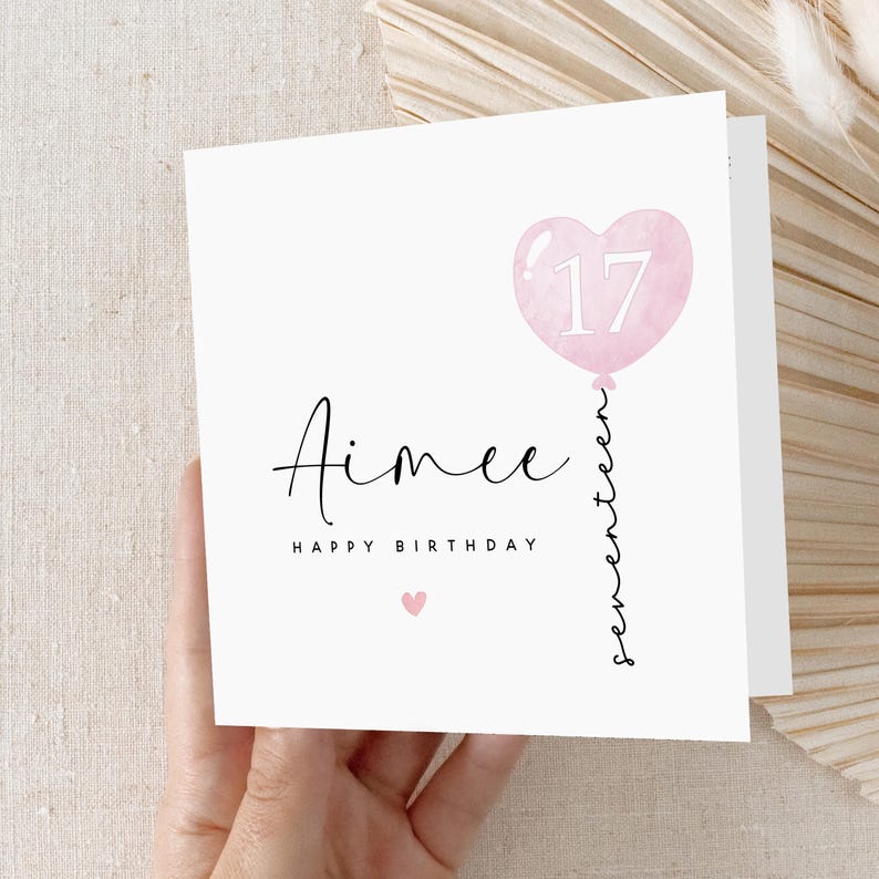 May include: A white card with a pink heart-shaped balloon with the number 17 inside. The text "Aimee Happy Birthday" is written in black cursive. The word "seventeen" is written vertically in cursive along the balloon string.