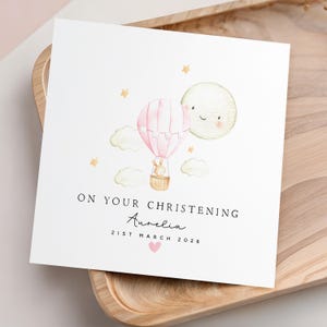 May include: A square white card with a watercolor illustration of a pink hot air balloon, a smiling moon, and stars. The text reads "ON YOUR CHRISTENING" with a name and date below, and a pink heart. The card is on a wooden tray.