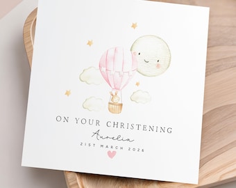 May include: A square white card with a watercolor illustration of a pink hot air balloon, a smiling moon, and stars. The text reads "ON YOUR CHRISTENING" with a name and date below, and a pink heart. The card is on a wooden tray.