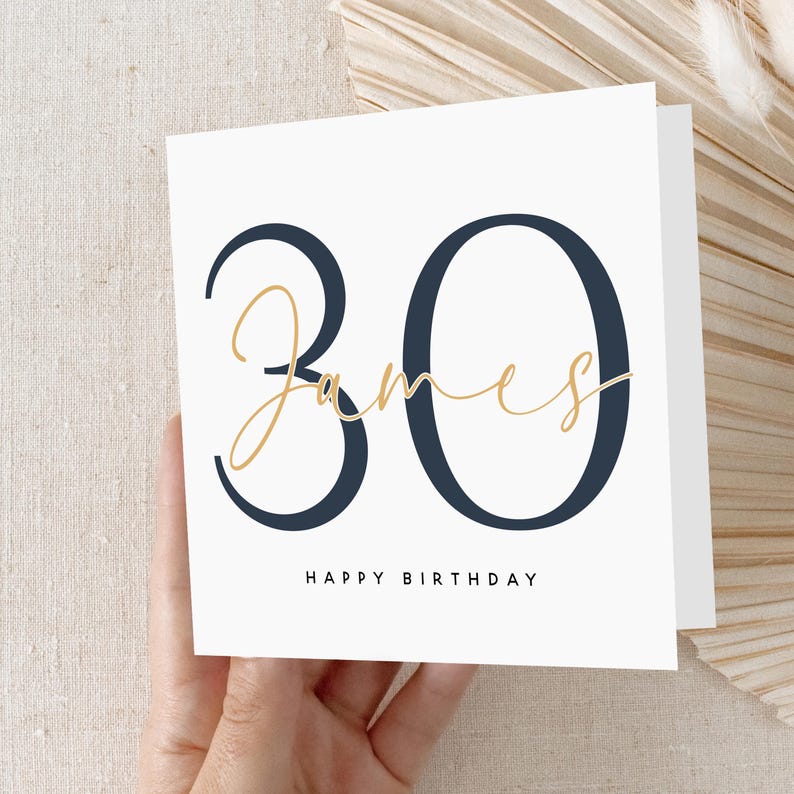 May include: A white card with a navy blue number "30" and the name "James" written in gold script. The text "Happy Birthday" is printed below the number.