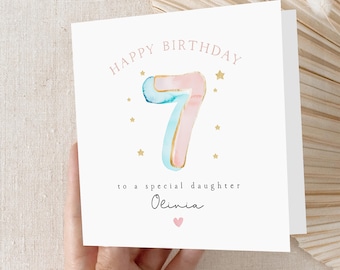 May include: A white birthday card with the text "HAPPY BIRTHDAY 7 to a special daughter Olivia" in pink and blue watercolor lettering. The number 7 is a watercolor design with gold accents, surrounded by gold stars and a pink heart.