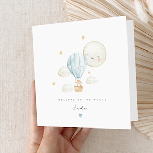 May include: A white card featuring a watercolour illustration of a blue hot air balloon with a small bear inside, flying alongside a smiling moon. The card reads "Welcome to the world Jude" with a small blue heart beneath the name.