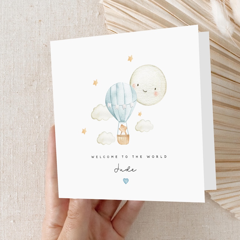 May include: A white card featuring a watercolour illustration of a blue hot air balloon with a small bear inside, flying alongside a smiling moon. The card reads "Welcome to the world Jude" with a small blue heart beneath the name.