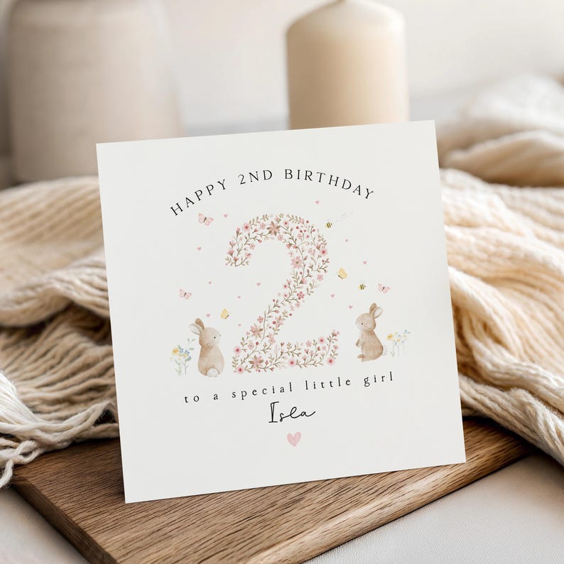 May include: A square birthday card with the text "HAPPY 2ND BIRTHDAY" at the top. The number "2" is filled with pink flowers. Two small bunnies and butterflies are on the card. The text "to a special little girl Isla" is below the number.
