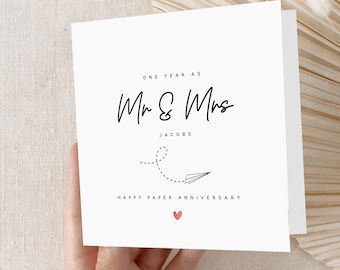 May include: A white anniversary card with black script that reads "ONE YEAR AS Mr & Mrs JACOBS". A paper airplane graphic and the words "HAPPY PAPER ANNIVERSARY" are below. A small red heart is at the bottom.
