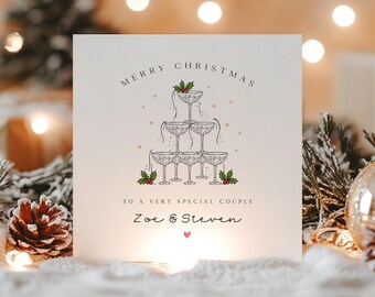 May include: A square white greeting card featuring a black line drawing of a champagne tower decorated with green holly leaves and red berries. The text reads Merry Christmas to a very special couple, Zoe & Steven, with a small pink heart below.