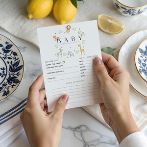 May include: A white baby shower prediction and advice card held by hands. The card features the word "BABY" with animal illustrations and sections for resemblance, birth predictions, and advice to parents.