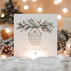 May include: A square greeting card featuring a central illustration of a silver Christmas ornament hanging from a green ribbon tied to snowy pine branches with red berries. The text reads Merry Christmas 2026 and James, Lucy & Jude with a small red heart.
