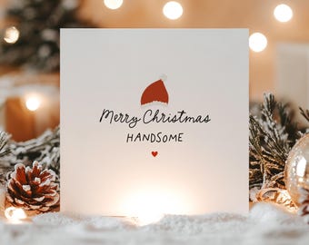 May include: A white Christmas card with the text "Merry Christmas Handsome" in black script, a red Santa hat illustration, and a small red heart. The card is set against a snowy backdrop with festive lights and pine cones.