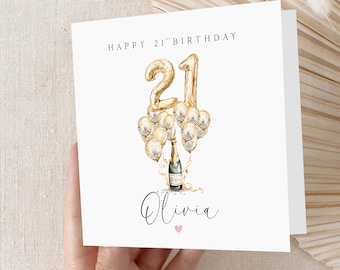 Op de afbeelding: Een witte verjaardagskaart met de tekst "HAPPY 21ST BIRTHAY" bovenaan. De kaart toont een gouden folieballon nummer 21, een champagnefles en ballonnen. De naam "Olivia" staat eronder in cursief, met een klein roze hartje.