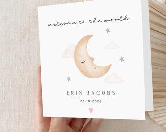 May include: A white card with a watercolor illustration of a crescent moon with a sleeping face. The moon is surrounded by white clouds and silver stars. The text "welcome to the world" is written in black cursive script. Below the moon is the name "ERIN JACOBS" and the date "02.10.2024" in black text. A small pink heart is at the bottom of the card.
