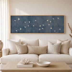 May include: A rectangular wall art piece with a blue background and a flock of white birds in flight. The artwork is framed in a light-colored wood and hangs above a white sofa with beige pillows. The room has a neutral color palette.