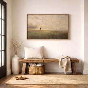 Puede incluir: Un cuadro de paisaje rectangular en un marco de madera clara cuelga sobre un banco de madera. La obra representa una figura solitaria en un campo vasto y brumoso bajo un cielo nublado. Un coj&iacute;n blanco, una cesta y una manta beige est&aacute;n presentes.