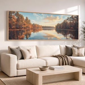 May include: A panoramic landscape painting in a light wood frame depicting a calm lake surrounded by dense autumn forests with orange and brown foliage under a soft blue sky with clouds, reflecting the trees and sky on the water surface.