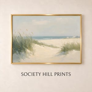 May include: A rectangular landscape painting in a thin gold frame depicting a sandy beach with tall green dune grass in the foreground, a calm blue ocean, and distant white buildings under a pale sky. The text SOCIETY HILL PRINTS appears below.