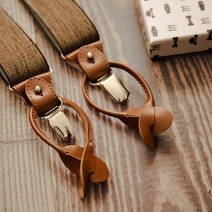 Può includere: Un paio di bretelle marroni con dettagli in pelle e clip in metallo argentato. Le bretelle sono su una superficie di legno.