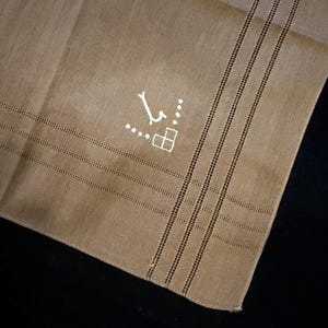 May include: A tan linen handkerchief featuring a white embroidered monogram letter J, accented by a small geometric square and dot pattern. The fabric displays a woven texture with parallel hemstitch detailing along the edges of the cloth.