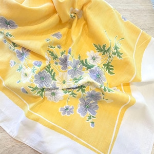 May include: A square yellow fabric cloth featuring a white border and a central floral print. The design displays clusters of light purple and white flowers with green leaves, arranged in a botanical pattern across the bright yellow background.