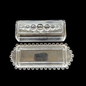 May include: Two-piece clear glass butter dish. The lid has a rectangular shape with a row of circular indentations. The base is rectangular with a beaded edge. The dish is designed to hold butter.
