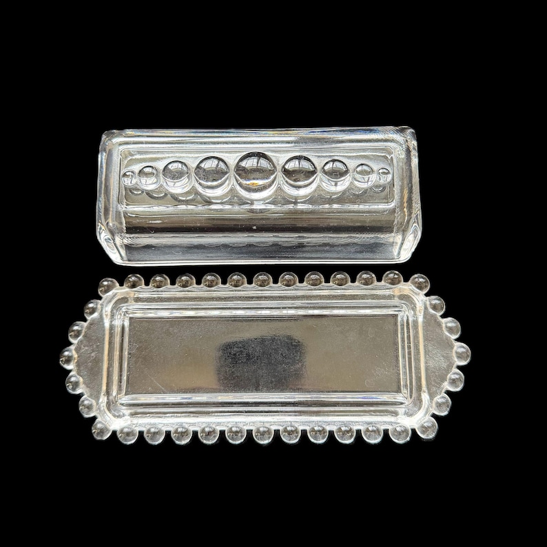 May include: Two-piece clear glass butter dish. The lid has a rectangular shape with a row of circular indentations. The base is rectangular with a beaded edge. The dish is designed to hold butter.
