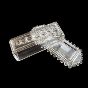 May include: Clear glass butter dish with a rectangular base and a lid. The lid and base have a decorative beaded edge. The lid has several circular indentations. The dish is made of clear glass.