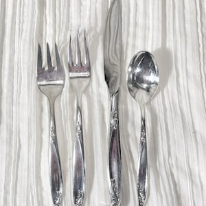 May include: A set of four silver-colored utensils: two forks, a knife, and a spoon. The utensils have a decorative floral pattern on the handles. The items are arranged on a white, textured surface.