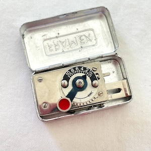 May include: A vintage light meter in a silver metal case. The meter has a black and silver dial with numbers and a blue pointer. A red button is on the side. The case is open, revealing the meter's components.