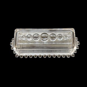 May include: Clear glass rectangular butter dish with a lid. The dish features a row of large, round, clear glass bubbles across the top and a row of smaller bubbles around the edge. The dish is on a black background.