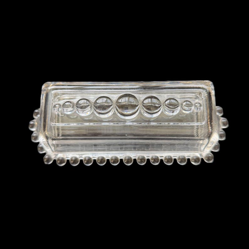 May include: Clear glass rectangular butter dish with a lid. The dish features a row of large, round, clear glass bubbles across the top and a row of smaller bubbles around the edge. The dish is on a black background.