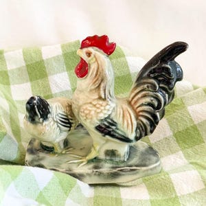 May include: A ceramic rooster and hen figurine. The rooster has a red comb and wattle, with a black and white tail. The hen is smaller and has black and white markings. The figurines are on a gray base, set against a green and white checkered background.