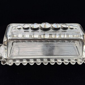 May include: Clear glass butter dish with a rectangular base and a lid. The lid is adorned with a row of round, raised glass accents. The base and lid have a beaded edge design. The dish is set against a black background.