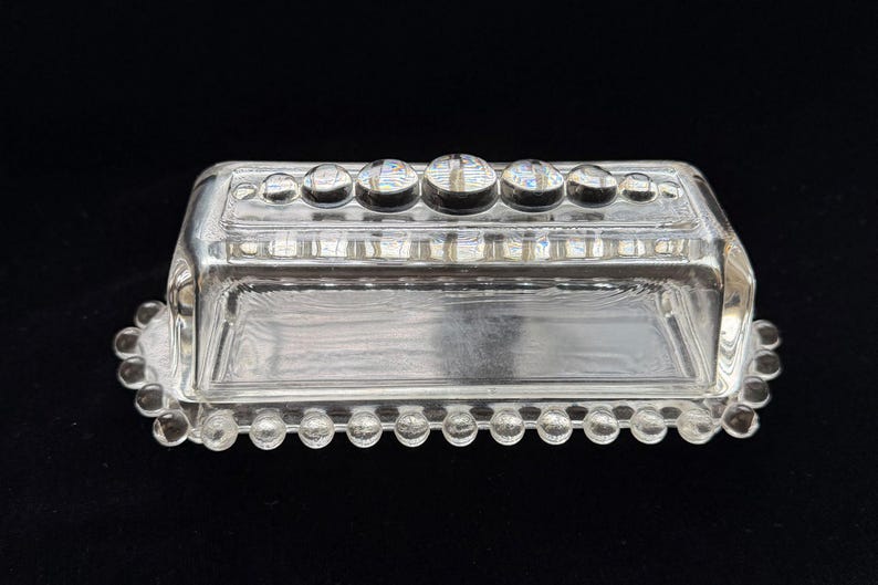 May include: Clear glass butter dish with a rectangular base and a lid. The lid is adorned with a row of round, raised glass accents. The base and lid have a beaded edge design. The dish is set against a black background.
