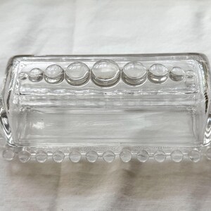 May include: A clear glass butter dish with a rectangular shape and a lid. The lid is adorned with a row of large, round, clear glass accents. The base and lid are edged with small, clear glass beads.