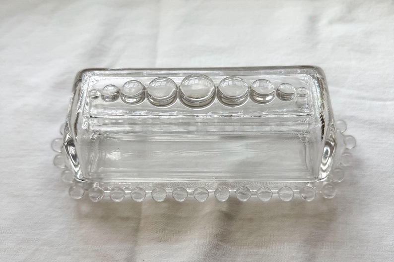 May include: A clear glass butter dish with a rectangular shape and a lid. The lid is adorned with a row of large, round, clear glass accents. The base and lid are edged with small, clear glass beads.