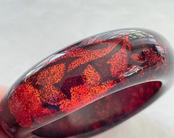May include: A red and black bangle bracelet. The bracelet is a deep red color with a glittery, textured pattern in shades of red and black. The bracelet is smooth and glossy.