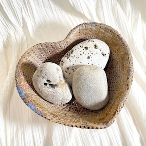 May include: A heart-shaped ceramic bowl with a textured, speckled surface in shades of brown, tan, and blue. The bowl contains three smooth, light-colored stones. The background is a white, textured fabric.