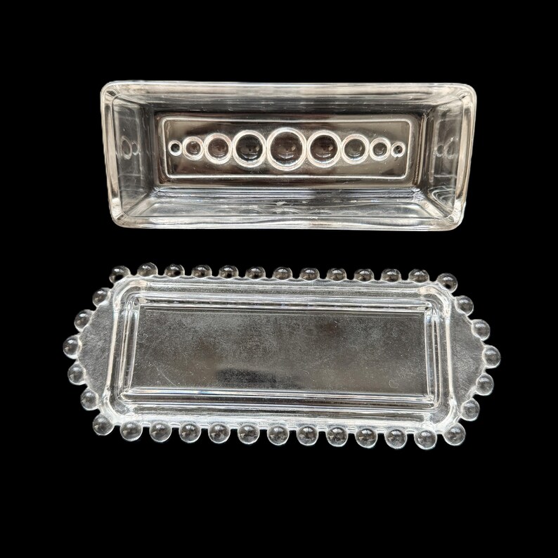 May include: Two clear glass rectangular trays. The top tray has a series of circular indentations. The bottom tray has a beaded edge. Both trays are on a black background.